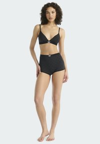 Kobieta z kręconymi włosami, nosząca czarny biustonosz Adidas i szorty z wysokim stanem, stoi boso na jasnym tle.