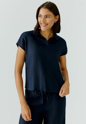 Femme souriante aux cheveux bruns mi-longs portant un haut à col bleu marine à manches courtes et un pantalon à cordon assorti.