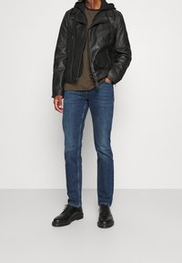 Chaqueta de cuero negra con bolsillos con cremallera, llevada sobre una camiseta de color verde olivo, combinada con jeans de mezclilla azul y zapatos negros de cordones.