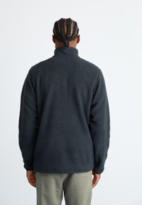 Person with braided hair wearing a dark gray fleece jacket and light gray pants, standing facing away against a plain light background.