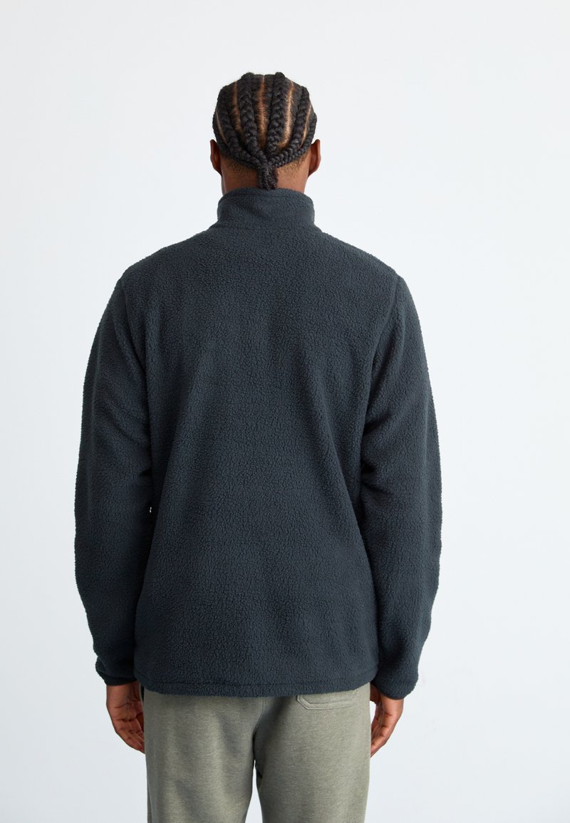 Person with braided hair wearing a dark gray fleece jacket and light gray pants, standing facing away against a plain light background.