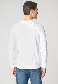 Maglione bianco a treccia con polsini e orlo a coste, caratterizzato da un collo rotondo e un motivo testurizzato, indossato con jeans blu.
