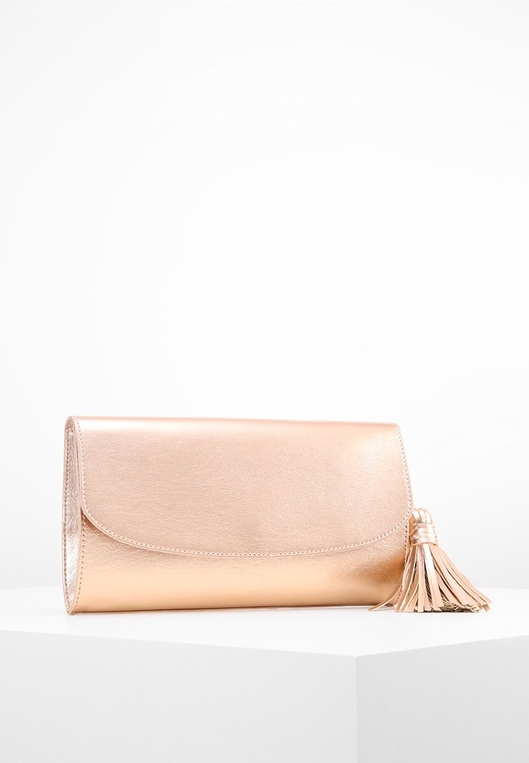 Lesklá kabelka v růžově zlaté barvě s klopou a velkým třásněm, vystavená na bílém podstavci na jednolitém bílém pozadí.