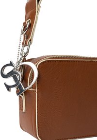 Sac en bandoulière en cuir marron avec une surface texturée, des accents blancs et un charme en argent présentant des initiales entrelacées. Fermeture éclair sur le dessus.