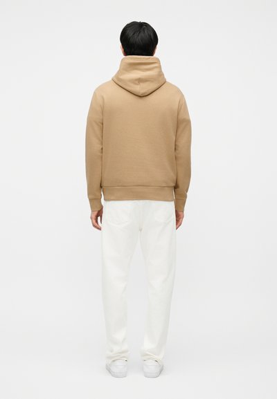 Άτομο στέκεται με την πλάτη γυρισμένη, φορώντας μπεζ φούτερ με κουκούλα, λευκό παντελόνι και λευκά παπούτσια, μπροστά σε ένα απλό φωτεινό φόντο.