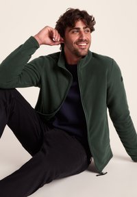 Green fleece jacket with a high collar, zip front, and black accents, worn over a navy shirt, paired with black pants.
