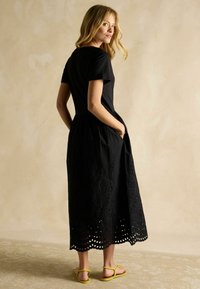 Melnas maksi kleitas ar īsām piedurknēm, kurai ir teksturēta izšūta apakšmala, pieguļošs augšdaļa un sānu kabatas. Akcentēta ar dzeltenām sandālēm.