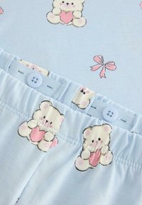 Ljusblå pyjamas-set med mönster av nallebjörnar som håller hjärtan, med en mjuk textur. Elastisk midja och knäppdetalj.