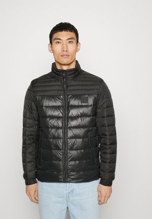 Giubbotto imbottito nero con collo alto, design trapuntato, texture liscia, chiusura con zip davanti e tasche laterali. Indossato sopra jeans azzurri chiari.
