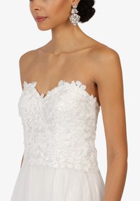 Robe blanche sans bretelles avec une superposition de dentelle florale et des accents perlés. Présente une jupe en tulle fluide et est accompagnée de grandes boucles d'oreilles ornées de bijoux.