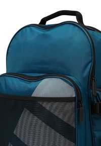 Tealtyg ryggsäck med rundad topp, med en meshnätskåp framtill och en svart dragkedja. Inuti finns ett tyg med en kontrasterande mönstring.