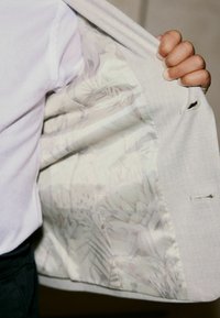Blazer gris clair avec un extérieur texturé et une doublure à motif floral, vu de l'intérieur, tenu ouvert par une main.