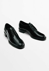 Miesi mustat nahkaiset puvuntossut, joissa on nauhat ja matalat korot, valkoisella taustalla.