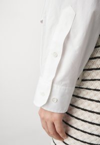 Manga de camisa blanca con puño puntiagudo, que cuenta con tres botones. El fondo muestra una falda estampada con rayas negras.