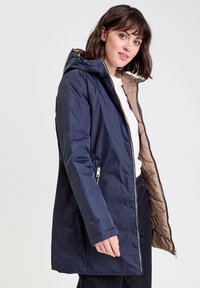 Femme portant un manteau à capuche bleu marine avec un doublure matelassée beige, une chemise blanche et un pantalon sombre, se tenant devant un arrière-plan clair.
