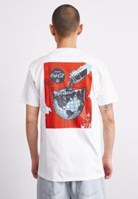Hvit bomulls T-skjorte med et fargerikt grafisk design av en klokke og en cola-flaske som helles over den, satt mot en lys rød bakgrunn.