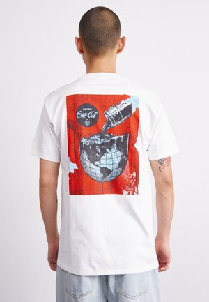 White cotton t-shirt featuring a colorful graphic design with a globe and a coke bottle pouring over it, set against a bright red background.