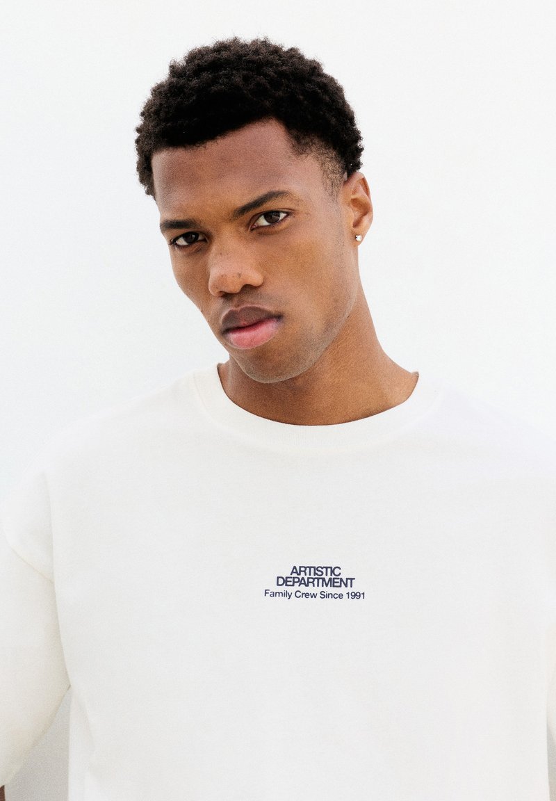 Jeune homme aux cheveux courts et bouclés portant un t-shirt blanc à col rond avec l'inscription « ARTISTIC DEPARTMENT Family Crew Since 1991 » sur la poitrine.