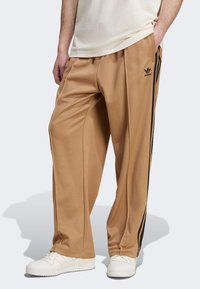 Pantalons de sport beiges avec des rayures latérales noires et un logo, fabriqués à partir d'un tissu lisse. Présentent une coupe ample et une taille élastique.