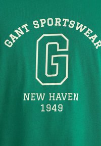 Camiseta de algodón verde con texto blanco: "GANT SPORTSWEAR," una gran "G," "NEW HAVEN," y "1949" en un diseño simple y audaz.