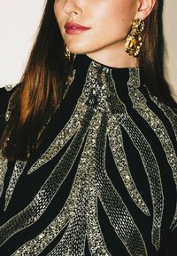 Top nero con dettagli di paillettes argentate a onde, abbinato a grandi orecchini dorati. Tessuto liscio con scollo alto.