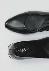 Décolleté in pelle nera con punta affusolata, texture liscia e soletta imbottita. Presenta il marchio "NEXT Motionflex" sulla soletta.