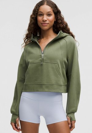 Jonge vrouw met krullend haar, gekleed in een olijfgroene cropped hoodie en witte sportieve shorts met hoge taille, staande tegen een effen achtergrond.