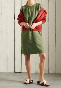 Olivenfarbenes Kleid mit rundem Ausschnitt, kurzen Ärmeln und lässiger Passform, kombiniert mit einer roten Bomberjacke. Schwarze Sandalen mit beigen Riemen.