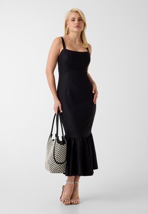 Femme debout dans une robe noire ajustée avec un ourlet volanté, tenant un sac cabas à motif noir et blanc, portant des sandales beiges à brides.