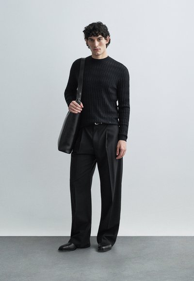 Μαύρο ριμπ πλεκτό πουλόβερ, χαλαρό μαύρο παντελόνι, μαύρο δερμάτινο messenger τσάντα, λείο υφής, στέκεται σε γκρι πάτωμα με φωτεινό φόντο.