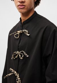 Juoda marškinė su stovinčiu apykaklė, papuošti trimis leopardų rašto juostelėmis ir subtilia tekstūra. Mygtukų užsegimas kaklo srityje.