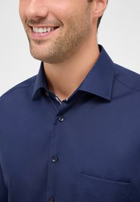 Camicia button-up blu navy con colletto strutturato, rivolta in avanti con taschino sul petto. Texture liscia e parte interna del colletto a righe in contrasto.