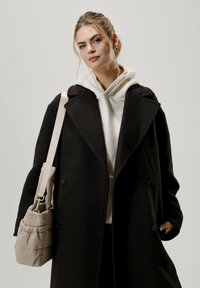 Musta villakangastakki, jossa on kaksirivinen nappitus, yllään valkoinen huppari. Mukana vaalea beige paisutettu laukku, jossa on olkahihna.