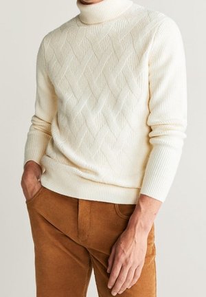 Cremefarbener Strickpullover mit hohem Kragen, verziert mit einem strukturierten Rautenmuster, kombiniert mit braunen Cordhosen.