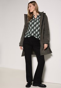 Langer olivgrüner Parka über einer gemusterten grün-weißen Bluse, kombiniert mit schwarzen Schlaghosen und schwarzen Stiefeletten. Sanfte Texturen sind zu bemerken.