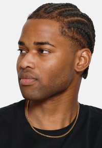 Primo piano di una persona con capelli intrecciati, che indossa una collana d'oro e una maglietta nera, evidenziando i tratti del viso e una texture della pelle liscia.