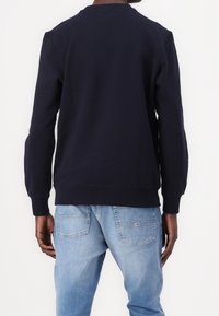 Uomo che indossa un felpa blu navy e jeans in denim azzurro, in piedi di spalle contro uno sfondo bianco uniforme.