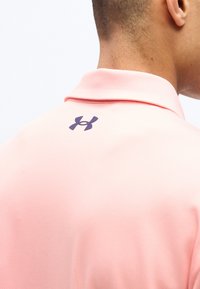 Κοντινή φωτογραφία της άνω πλάτης ενός ατόμου που φοράει ροζ πουκάμισο με γιακά, με ένα μαύρο λογότυπο Under Armour κάτω από τον γιακά.