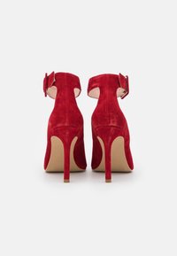 Chaussures à talons hauts en suède rouge avec une sangle à la cheville dotée d'une boucle et de découpes. Texture lisse et design à bout rond.