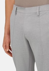 Gros plan sur un pantalon gris clair texturé avec passants de ceinture, porté par une personne la main détendue le long du corps.