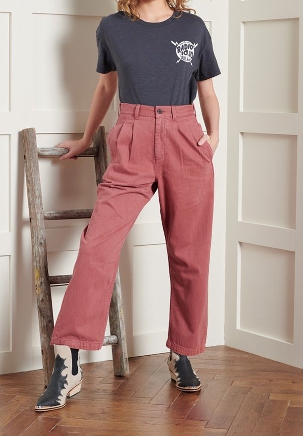 Personne portant un t-shirt graphique noir, un pantalon rose ample taille haute et des bottines bicolores, debout à côté d’une échelle en bois à l’intérieur.