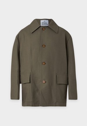 Jeune homme portant une veste boutonnée vert olive, un pantalon beige, des chaussures noires, debout devant un fond blanc uni.