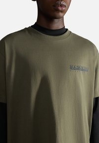 T-shirt in cotone verde oliva con colletto a giro e maniche corte. Presenta un logo grigio stampato "Napapijri" sul lato sinistro. Texture liscia.