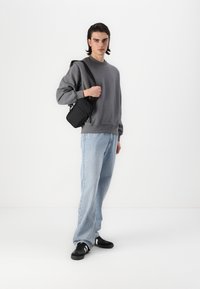 Felpa grigia, jeans azzurri chiari, sneakers nere e una piccola borsa a spalla nera. Sfondo neutro, illuminazione naturale.