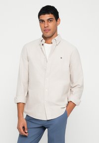 Svetlo béžová košeľa na gombíky z ľanovej zmesi, so štandardným golierom a vyhrnutými rukávmi, doplnená modrými nohavicami, ktoré majú malé logo na hrudníku.