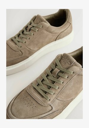 Pereche de adidași din piele intoarsă taupe cu șireturi verde măsliniu și talpă albă de cauciuc pe un fundal deschis.