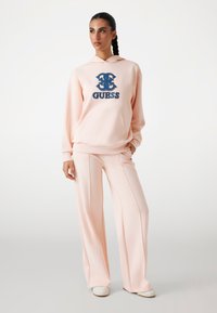 Sudadera rosa claro con un logo "GUESS" en navy; combinada con pantalones de pierna ancha que cuentan con bolsillos laterales y una textura suave.