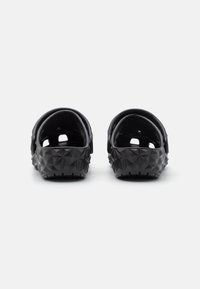 Sabots en caoutchouc noir avec un motif géométrique texturé sur les côtés, ouvert à l'arrière et avec une sangle ajustable. Intérieur lisse et semelle antidérapante.