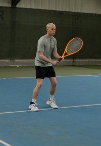 Tenisista w jasnoszarej koszulce i czarnych szortach, trzymający pomarańczową rakietę. Znajduje się na niebieskim korcie halowym z zielonym tłem.