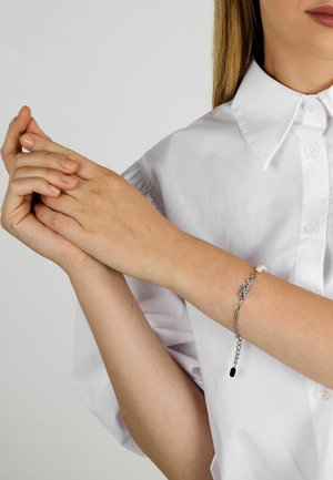 Vrouw in wit shirt die haar linkerpols vasthoudt, met een zilveren kettingarmband met een kleine vlindertag en een pareldetail.